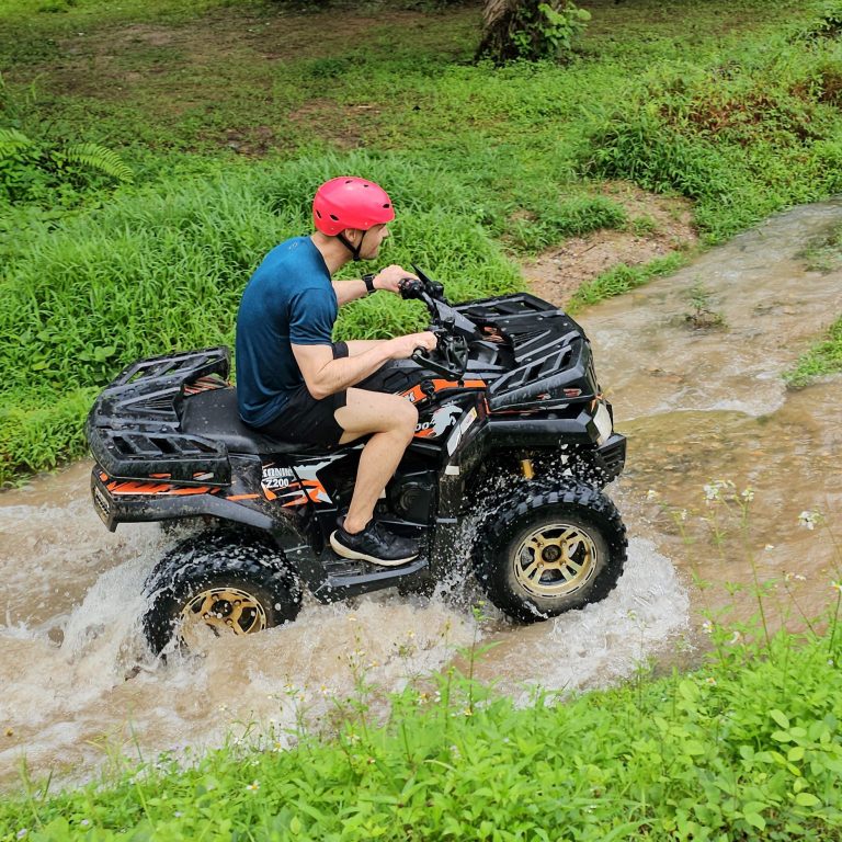 ATV rider in a red helmet driving through a shallow stream in the countryside